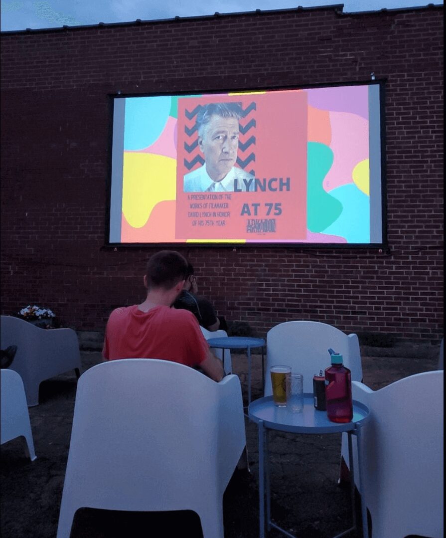 Outdoor event showing a man watching a screen displaying a tribute to David Lynch at 75.
